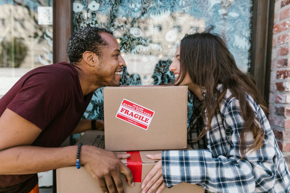 A man and woman are outside a property, engaging in a home relocation activity by exchanging boxes. The man, of African descent with curly hair, is smiling and leaning forward, holding a cardboard box