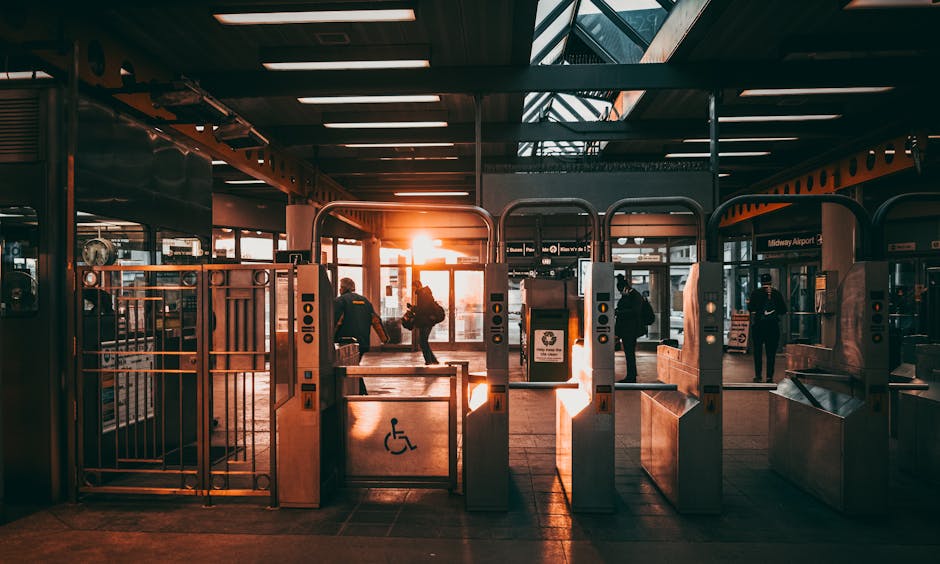 Inside Stratford Station during sunset, a row of automated ticket gates and turnstiles with card readers and barrier arms are visible, leading toward the station platform. Several commuters with luggage and backpacks are walking through the gates, some carrying or wheeling suitcases and bags, heading towards the exit or platform. The station features a modern, spacious interior with a high ceiling and large glass windows allowing the warm glow of the setting sun to illuminate the space. The platform area beyond the gates shows benches and signage, including a 'Midway Airport' sign, indicating an airport transfer service. The advanced barriers include a designated space for disabled access, as seen with the wheelchair-friendly gate. This scene captures a typical busy urban transport hub, where furniture and boxes could be loaded onto vehicles for a home relocation—an environment familiar to [COMPANY_NAME] when coordinating furniture transport and packing during house removals involving tight access, as outlined in the Stratford Station removals guide for tight access, STRATFORD.