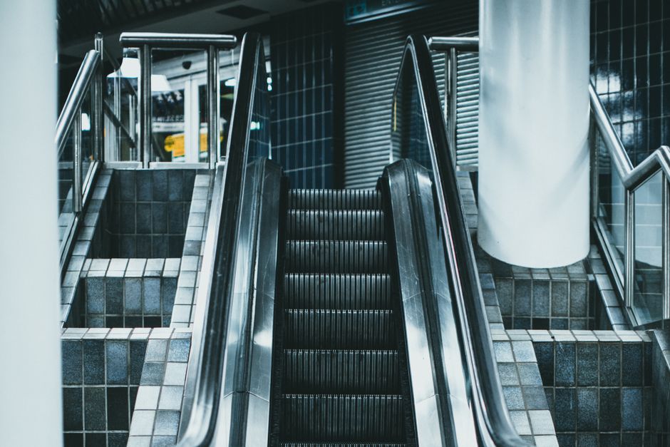 A set of escalators inside a building, with the central escalator in black metal steps and handrails on either side, leading upwards. To the left, there is a small platform with a dark tiled surface, bordered by a glass and metal railing, while to the right, a white cylindrical column is visible near the escalator. The surroundings include dark tiled walls and a metallic shutter or rolling door positioned behind the escalator, suggesting an entrance or exit area typical of a transport hub such as a train station. The lighting appears to be artificial, highlighting the reflective surfaces of the escalator and the glossy tiles. This scene depicts interior movement and infrastructure relevant to house removals, conveying the process of furniture transport and logistics within urban environments, as handled by a professional moving company like Man with Van Stratford.